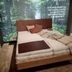 Bett mit Matratze und Leinenbezug vor bewaldeter Fensterfront