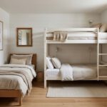 Ein helles, hochwertig eingerichtetes kleines Schlafzimmer im skandinavischen Stil in einer Ferienwohnung mit einem Etagenbett und einem Einzelbett zur Maximierung der Schlafplätze. An der Wand hängt ein gerahmtes Poster mit dem Titel „Airbnb Schlafplätze maximieren: Mehr Buchungen, mehr Umsatz“