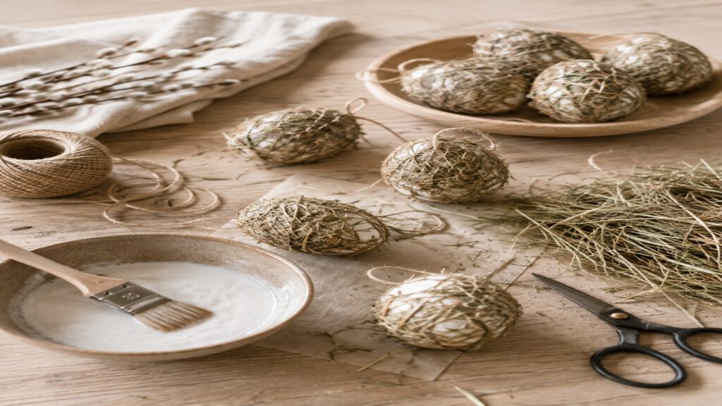 Basteltisch mit Ostereiern aus Heu, Kleisterschale, Pinsel und Naturbindfaden